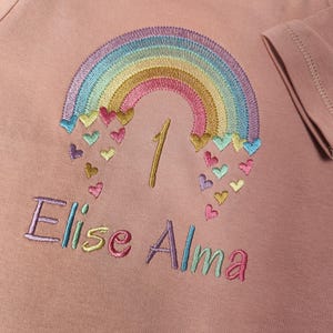 Puede incluir: Una camiseta de manga larga en color rosa empolvado con un arco&iacute;ris bordado del que caen peque&ntilde;os corazones. El n&uacute;mero 1 est&aacute; cosido con hilo dorado bajo el arco&iacute;ris y el nombre Elise Alma est&aacute; bordado en letras de colores debajo.