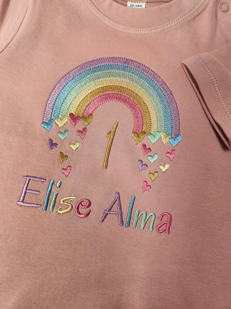 Puede incluir: Una camiseta de manga larga en color rosa empolvado con un arco&iacute;ris bordado del que caen peque&ntilde;os corazones. El n&uacute;mero 1 est&aacute; cosido con hilo dorado bajo el arco&iacute;ris y el nombre Elise Alma est&aacute; bordado en letras de colores debajo.