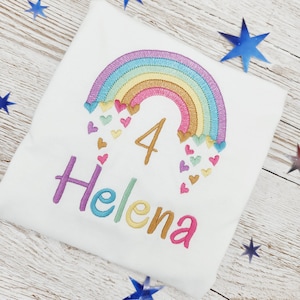 Puede incluir: Una camiseta blanca con un dise&ntilde;o bordado de un arco&iacute;ris multicolor con peque&ntilde;os corazones cayendo de sus extremos. El n&uacute;mero 4 est&aacute; cosido en hilo dorado debajo del arco&iacute;ris y el nombre Helena est&aacute; bordado en letras multicolores.