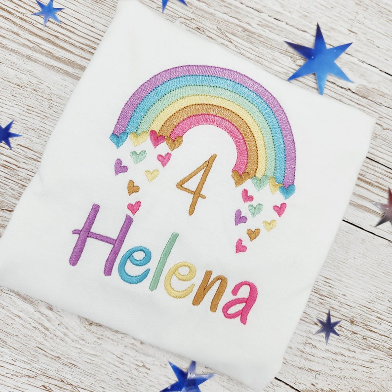 Puede incluir: Una camiseta blanca con un dise&ntilde;o bordado de un arco&iacute;ris multicolor con peque&ntilde;os corazones cayendo de sus extremos. El n&uacute;mero 4 est&aacute; cosido en hilo dorado debajo del arco&iacute;ris y el nombre Helena est&aacute; bordado en letras multicolores.