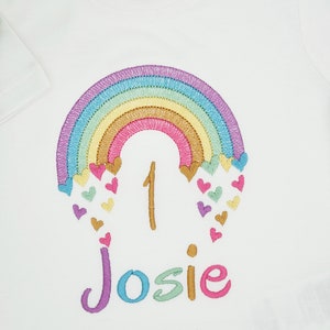 Puede incluir: Una camiseta blanca de manga corta con un dise&ntilde;o de arco&iacute;ris bordado con peque&ntilde;os corazones de colores, el n&uacute;mero 1 y el nombre Josie en hilo multicolor. Una etiqueta blanca en el cuello muestra el texto 12-18m y 76-86cm.