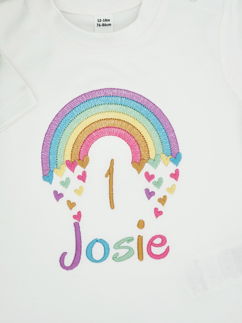 Puede incluir: Una camiseta blanca de manga corta con un dise&ntilde;o de arco&iacute;ris bordado con peque&ntilde;os corazones de colores, el n&uacute;mero 1 y el nombre Josie en hilo multicolor. Una etiqueta blanca en el cuello muestra el texto 12-18m y 76-86cm.
