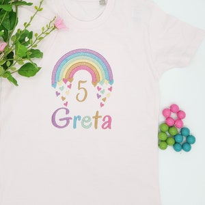 Puede incluir: Una camiseta de manga corta de color rosa claro con un dise&ntilde;o bordado de un arco&iacute;ris en tonos pastel con peque&ntilde;os corazones y el n&uacute;mero 5 en dorado. Debajo del arco&iacute;ris, el nombre Greta est&aacute; bordado en letras multicolores en el frente.