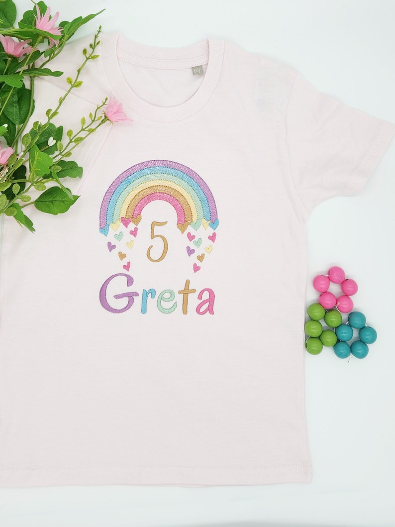 Puede incluir: Una camiseta de manga corta de color rosa claro con un dise&ntilde;o bordado de un arco&iacute;ris en tonos pastel con peque&ntilde;os corazones y el n&uacute;mero 5 en dorado. Debajo del arco&iacute;ris, el nombre Greta est&aacute; bordado en letras multicolores en el frente.