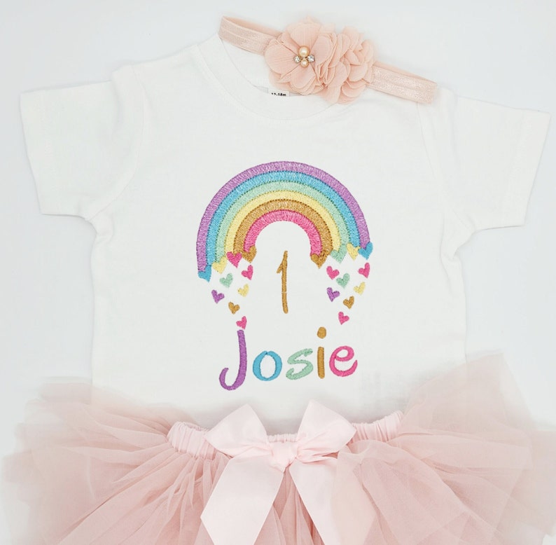 Puede incluir: Una camiseta blanca de manga corta con un arco&iacute;ris bordado de rayas doradas, azules y moradas, peque&ntilde;os corazones de colores, el n&uacute;mero 1 y el nombre Josie en hilo multicolor. Incluye una falda de tul rosa y una diadema a juego.