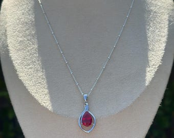 May include: A silver necklace with a teardrop-shaped, red gemstone pendant. The pendant is encased in a silver swirl design, and the chain is delicate. The necklace is displayed on a mannequin.