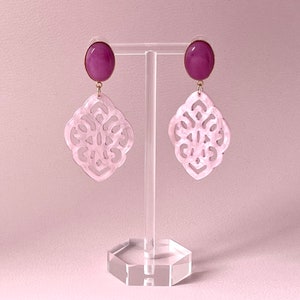Könnte beinhalten: Ein Paar Hängeohrringe mit einem rosa-weißen Design. Die Ohrringe haben einen runden, violetten Stein oben und ein weißes, filigranes, rautenförmiges Design darunter.