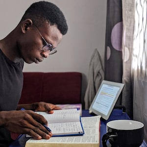 Puede incluir: Una persona con gafas se sienta en un escritorio con una superficie azul, leyendo un libro grande abierto. Un bolígrafo azul está en su mano, y una taza de café negra se encuentra en el escritorio a la derecha. Una tableta está visible en el fondo.