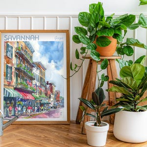 May include: A watercolor painting of a street scene in Savannah, Georgia. The painting depicts a row of colorful buildings with balconies and storefronts. The street is lined with trees and people walking by. The painting is framed in a wooden frame and is hanging on a white wall.