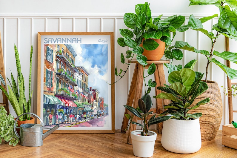 May include: A watercolor painting of a street scene in Savannah, Georgia. The painting depicts a row of colorful buildings with balconies and storefronts. The street is lined with trees and people walking by. The painting is framed in a wooden frame and is hanging on a white wall.