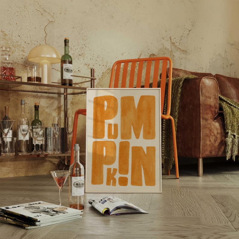 May include: A framed art print with the word "PUMPKIN" in orange block letters. The print is propped against an orange metal chair. A bar cart with bottles and glasses, a leather couch, and a magazine are also in the image.