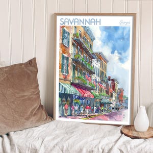 May include: A watercolor painting of a street scene in Savannah, Georgia. The painting depicts a row of colorful buildings with balconies and awnings. There are people walking on the street and trees lining the sidewalk. The painting is framed in a wooden frame.