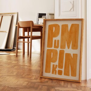 May include: A framed art print with the word "PUMPKIN" in large, orange block letters. The artwork is displayed on a wooden floor, leaning against a white wall. The room has a minimalist, modern aesthetic with natural light.