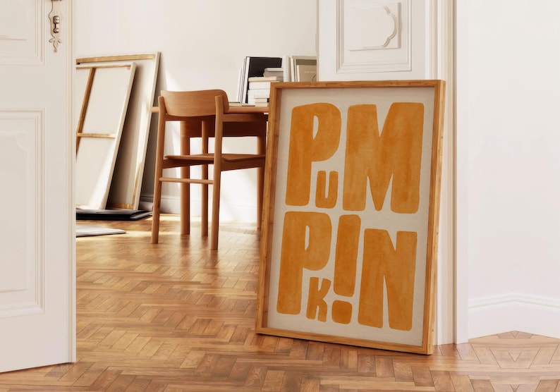 May include: A framed art print with the word "PUMPKIN" in large, orange block letters. The artwork is displayed on a wooden floor, leaning against a white wall. The room has a minimalist, modern aesthetic with natural light.