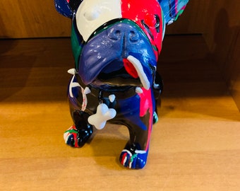 May include: A colorful ceramic French bulldog figurine. The dog's face is split with white and red paint, while the body is painted with blue, black, green, and red. A white bone hangs from its neck.