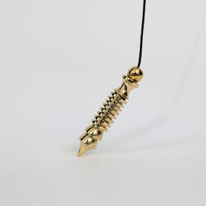 May include: A gold-colored metal pendulum with a ribbed, cylindrical body and a pointed tip, suspended from a thin black cord. The object features a smooth, spherical top piece and a series of horizontal ridges along its central shaft.