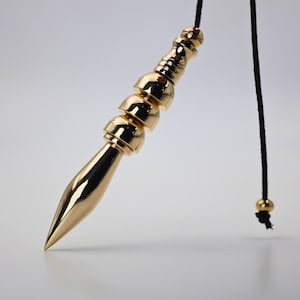 Puede incluir: Un péndulo dorado con punta y una serie de segmentos redondeados. Cuelga de un cordón negro con una cuenta dorada. El diseño es elegante y moderno, adecuado para la adivinación o la decoración.