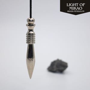 Könnte beinhalten: Ein silberfarbenes Messingpendel, das an einer schwarzen Schnur hängt. Das Pendel hat eine Spitze und einen zylindrischen Körper mit gerippten Details. Der Text "LIGHT OF MIKAO BRASS PENDULUM" befindet sich oben rechts.