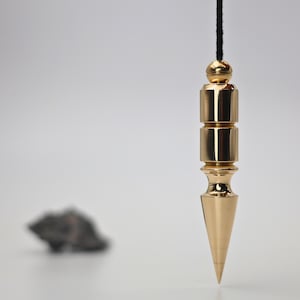 May include: A gold-colored pendulum suspended by a black cord. The pendulum has a cylindrical body with a pointed tip. The background is a soft, neutral white, with a blurred dark gray rock in the distance.