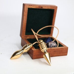 May include: Two gold-colored pendulums with pointed tips and attached cords rest near an open wooden box. The box is lined with green fabric and contains several small, polished stones. The pendulums are displayed on a white surface.