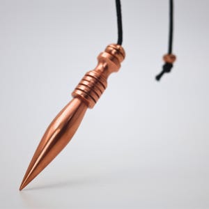 May include: A copper-coloured pendulum with a pointed tip and a ridged handle, suspended by a black cord. The design is simple and elegant, suitable for divination or decorative purposes.