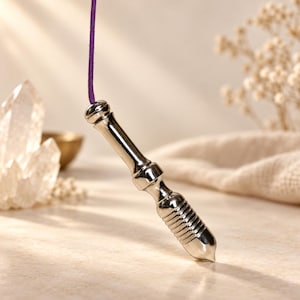 May include: A polished silver-toned metal pendulum with a smooth, tapered upper section and a ribbed, cylindrical lower body. The pendulum is suspended from a thin, vibrant purple cord, showcasing a reflective, metallic finish throughout its design.