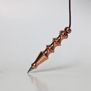 May include: A copper-coloured pendulum with a pointed tip and decorative, ridged design. The pendulum is suspended by a brown cord. The background is a plain white surface.