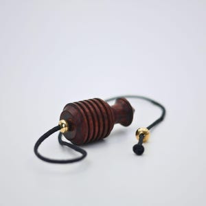 May include: A dark brown wooden pendulum with a black cord and gold-colored accents. The pendulum has a cylindrical body with horizontal grooves and a rounded top. The cord is attached to a gold-colored bead.