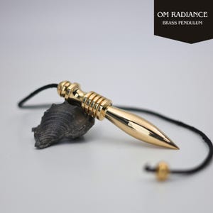 Può includere: Un pendolo in ottone lucido con punta e cordino nero. Il pendolo è appoggiato su una pietra grigio scuro. Il testo "OM RADIANCE BRASS PENDULUM" si trova in alto a destra.