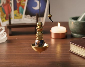 Könnte beinhalten: Ein Messingpendel mit einer blauen und braunen Steinkugel, die an einer schwarzen Schnur hängt. Das Pendel befindet sich vor Tarotkarten, einer brennenden Kerze und einem Mörser und Stößel, was auf ein spirituelles oder divinatorisches Thema hindeutet.