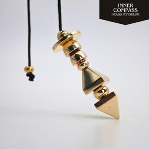 Op de afbeelding: Een goudkleurige messing slinger met een geometrisch ontwerp, opgehangen aan een zwart koord. De slinger heeft een reeks vormen, waaronder een schijf, cilinders, bollen en een piramide. De tekst "INNER COMPASS BRASS PENDULUM" staat rechtsboven.