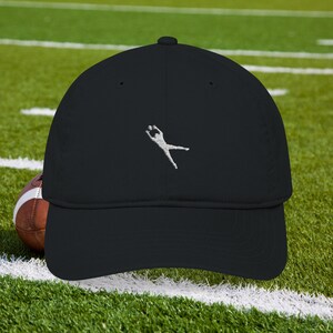 May include: Black baseball cap with a white embroidered figure of a person catching a football. The cap is made of a soft material and has a curved brim. A brown and white football is visible in the background.