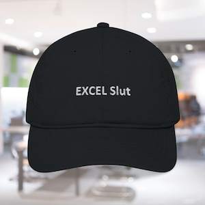 May include: Black baseball cap with the embroidered text "EXCEL Slut" in white. The cap has a curved brim and a rounded crown. The background is blurred, suggesting an indoor setting.