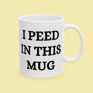 Może przedstawiać: Biały ceramiczny kubek z zakrzywionym uchwytem. Kubek ma czarny napis "I PEED IN THIS MUG" pogrubioną czcionką bezszeryfową. Kubek jest ustawiony na jasnym żółtym tle.