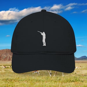 May include: Black baseball cap with a white embroidered silhouette of a person aiming a rifle. The cap has a curved brim and a structured fit. The background shows a grassy field, mountains, and a blue sky with clouds.