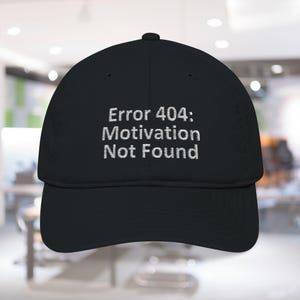 May include: Black baseball cap with the embroidered text "Error 404: Motivation Not Found" in white. The cap has a curved brim and a structured crown. The background is blurred, suggesting an indoor setting.