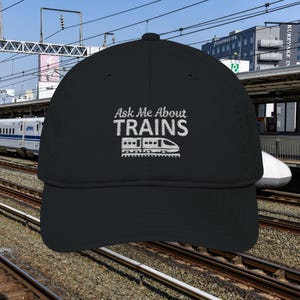May include: Black baseball cap with the embroidered text "Ask Me About TRAINS" and a train graphic in white. The cap is set against a background of train tracks and a train station.
