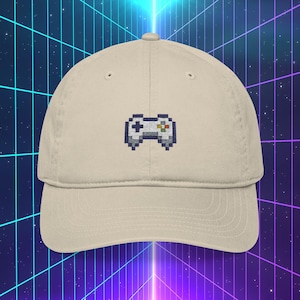 May include: A beige baseball cap featuring an embroidered pixelated video game controller design. The controller is in shades of gray, blue, red, and green. The cap has a curved brim and a classic six-panel construction.