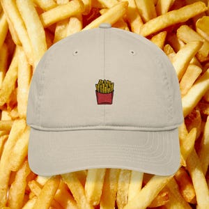 May include: A beige baseball cap featuring an embroidered image of a red carton of french fries. The cap is set against a background of golden french fries. The cap has a curved brim and a classic six-panel design.