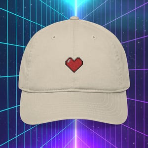 May include: A beige baseball cap featuring a pixelated red heart design embroidered on the front. The cap has a curved brim and a classic six-panel construction. The hat is made of a soft, breathable fabric.