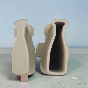May include: Two beige, abstract, vase-shaped objects are displayed against a light blue background. The objects have a smooth, matte finish and a unique, modern design. One object is open, revealing the interior, while the other is closed.