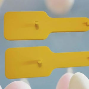 May include: Two yellow popsicle molds with a rectangular base and a handle. Each mold has a small peg in the center of the base and a small hole at the end of the handle. The molds are set against a blurred background of pastel colors.