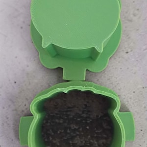 May include: A green, 3D-printed cauldron-shaped container with a lid. The cauldron is open, revealing a dark, granular substance inside. The lid is also green and shaped to fit the cauldron. The design is reminiscent of a witch's cauldron.