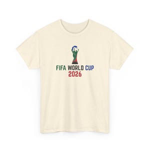 May include: A cream-colored short-sleeve t-shirt with a speckled texture. The center of the chest features a graphic of the FIFA World Cup trophy in green, black, and blue, with the text FIFA WORLD CUP in green and blue, and 2026 in red below.