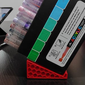 May include: A black marker box with a variety of acrylic markers in shades of purple, pink, and clear. The box is propped up on a red, honeycomb-patterned stand. The box has color-coded tabs and instructions for use.