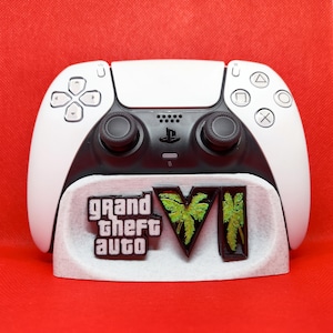 Pode incluir: Controlo PlayStation 5 branco e preto sobre um suporte branco. O suporte apresenta o logótipo "Grand Theft Auto VI" em preto, verde e branco. O fundo é vermelho liso.