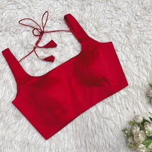 Puede incluir: Una blusa roja sin mangas con escote cuadrado y tirantes anchos. La parte posterior de la prenda incluye finas cintas de tela roja con borlas a juego para el cierre, proporcionando un ajuste seguro para este top corto de color sólido.