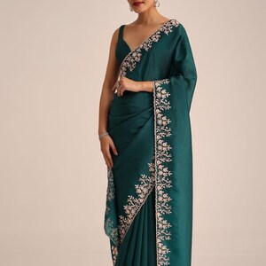May include: A deep emerald green saree featuring a sleeveless blouse and a matching drape. The fabric has a subtle textured finish, and the borders are adorned with intricate light pink floral embroidery and delicate sequin detailing throughout.