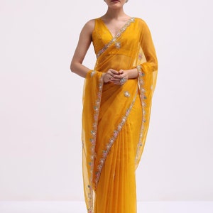 May include: A vibrant mustard yellow saree featuring a sleeveless blouse with a deep V-neckline. The sheer fabric of the saree is adorned with a delicate floral embroidery border in shades of pink, blue, and white, accented with small silver sequins.