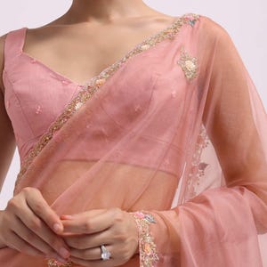 May include: A blush pink saree with detailed floral embroidery on the borders and sleeves. The sheer fabric drapes elegantly, paired with a matching blouse. Delicate embellishments add a touch of glamour to this formal wear.
